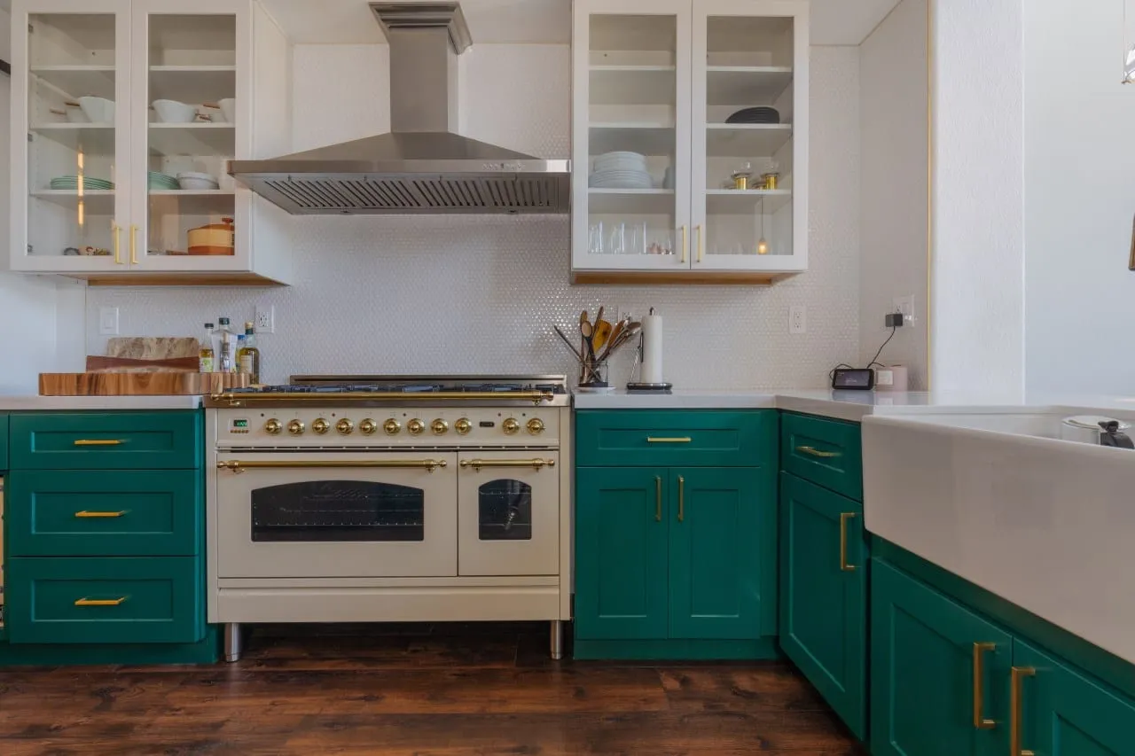 Cream range with brass trim set under a stainless chimney hood between emerald cabinets