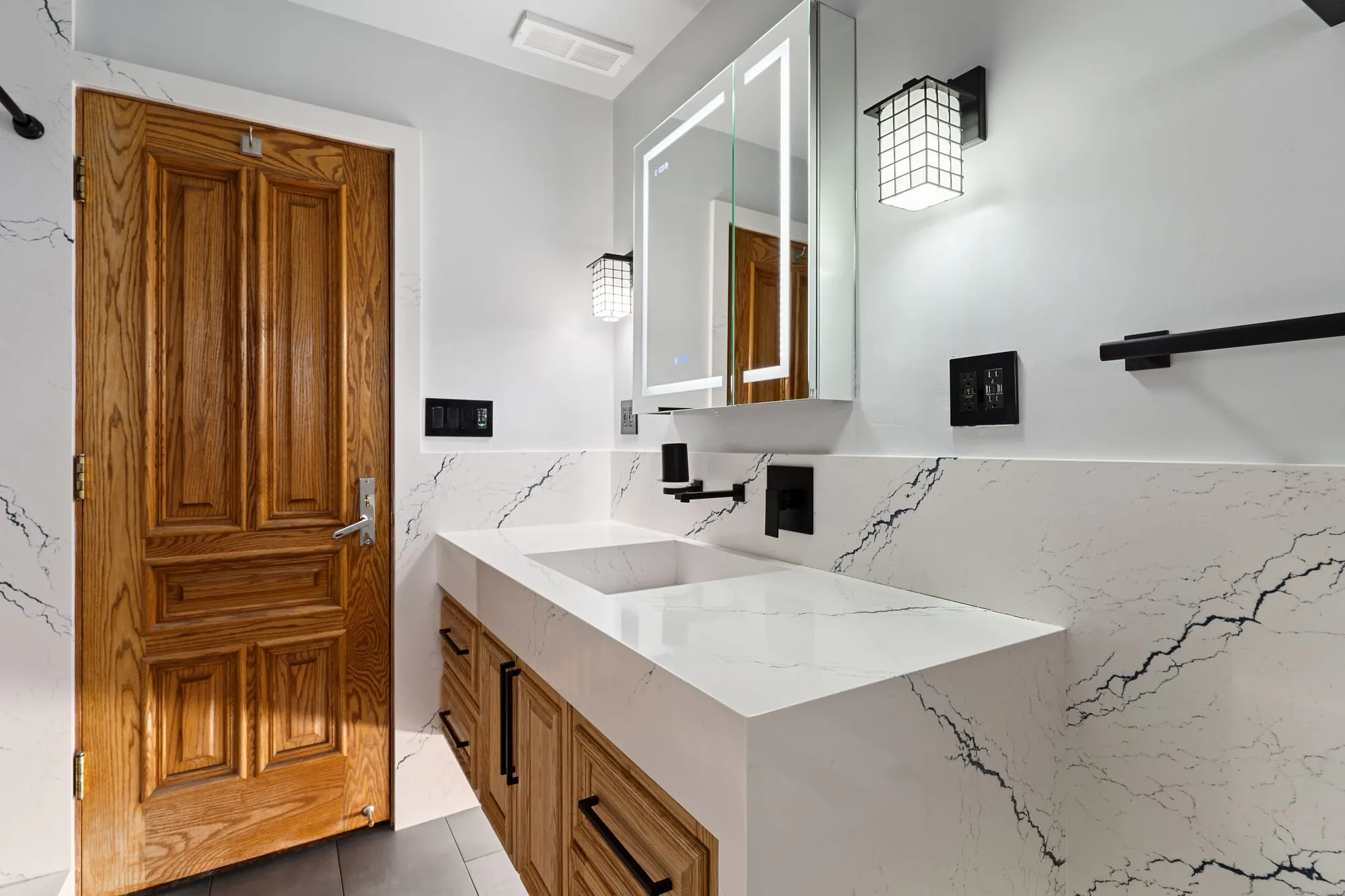 Bathroom after remodel, marble-look slab walls with a backlit mirror above an oak vanity