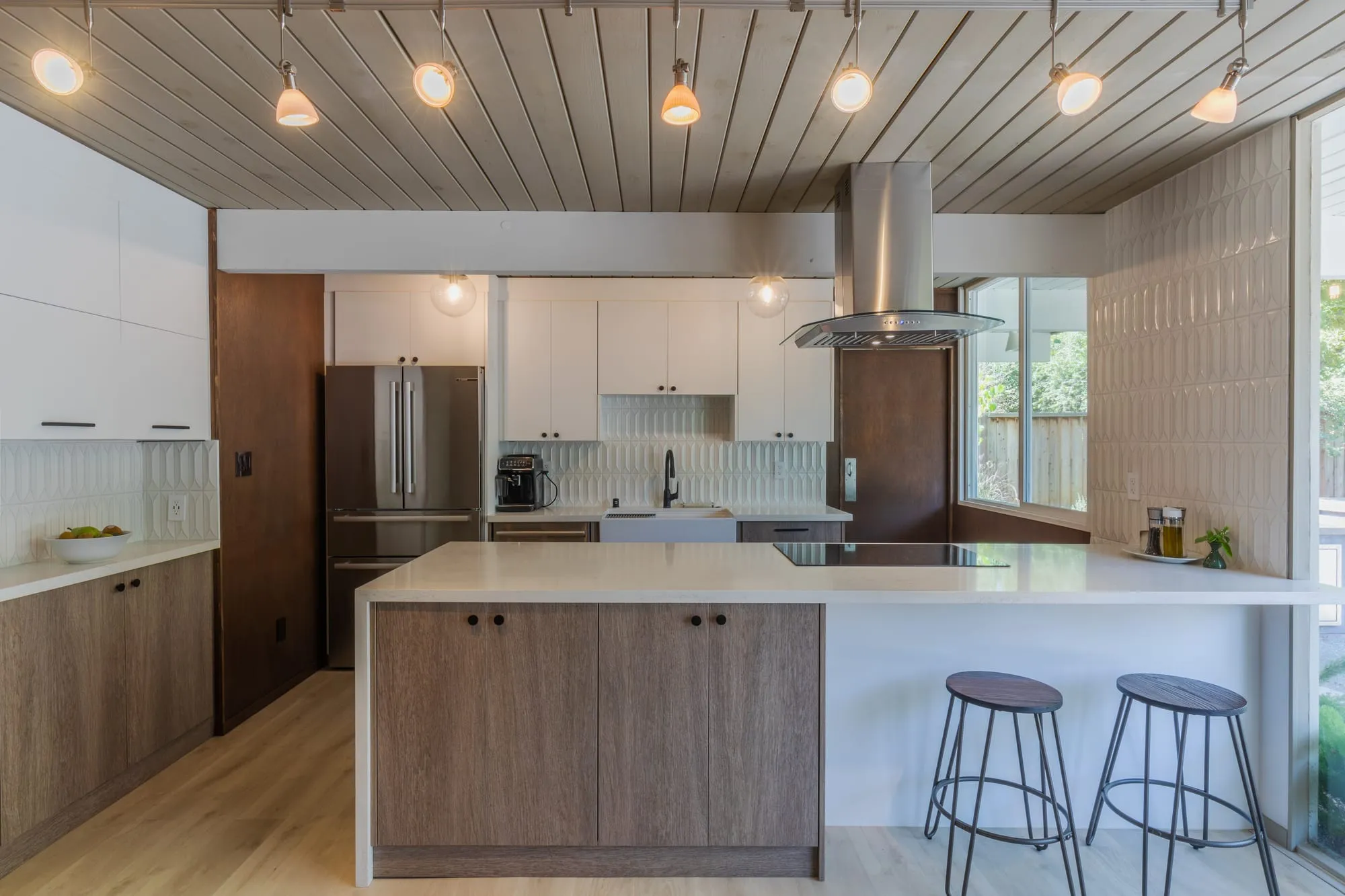 Kitchen after remodel, rift-oak cabinetry and white uppers around a quartz island under a plank ceiling