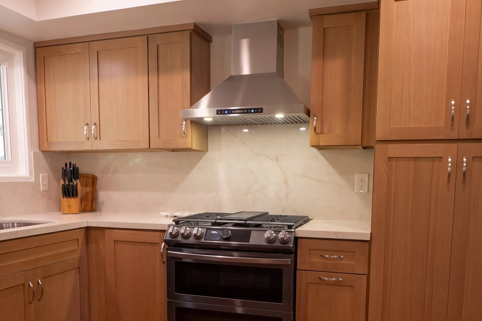 Kitchen after remodel, wood shaker cabinetry under a full-height quartz backsplash with a stainless chimney hood